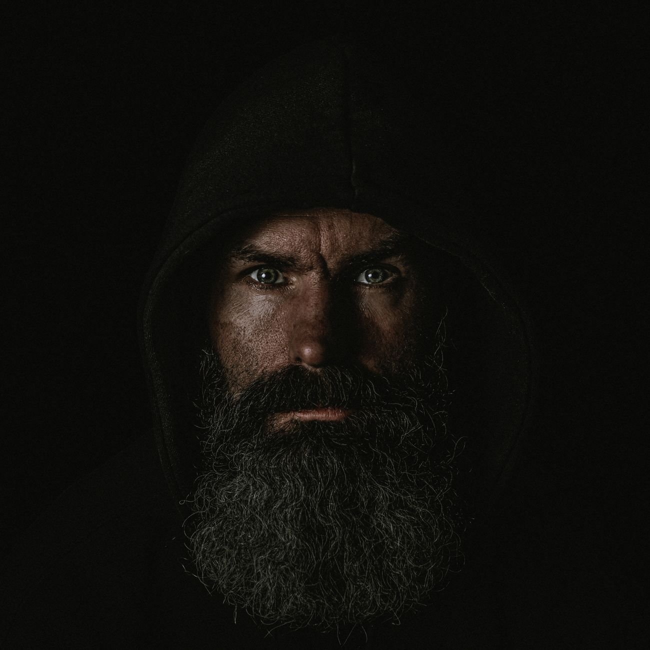 Dramatic portrait of a bearded man wearing a hoodie against a black background.