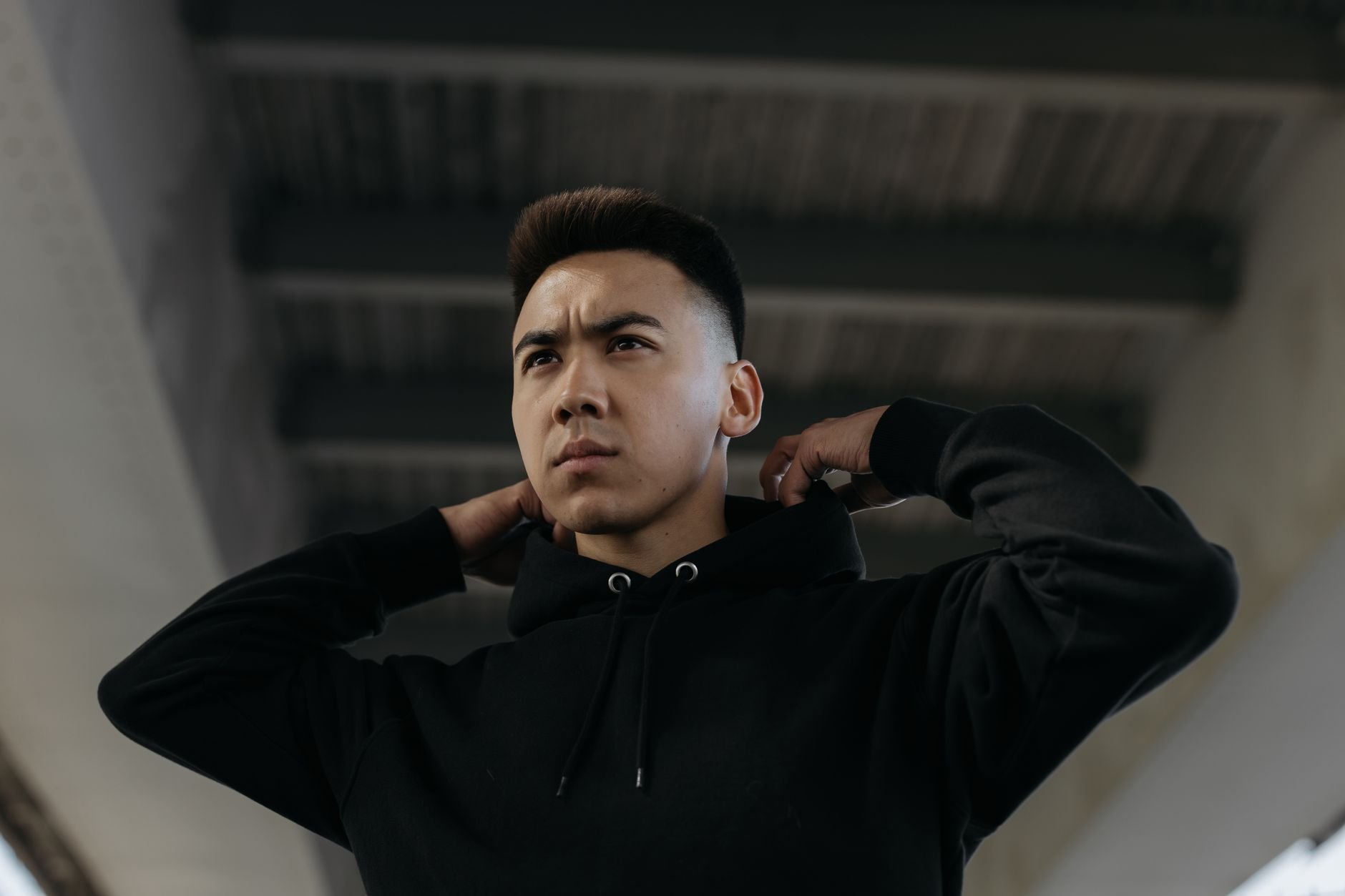 A determined young man wearing a black hoodie adjusts his hood under an urban bridge.